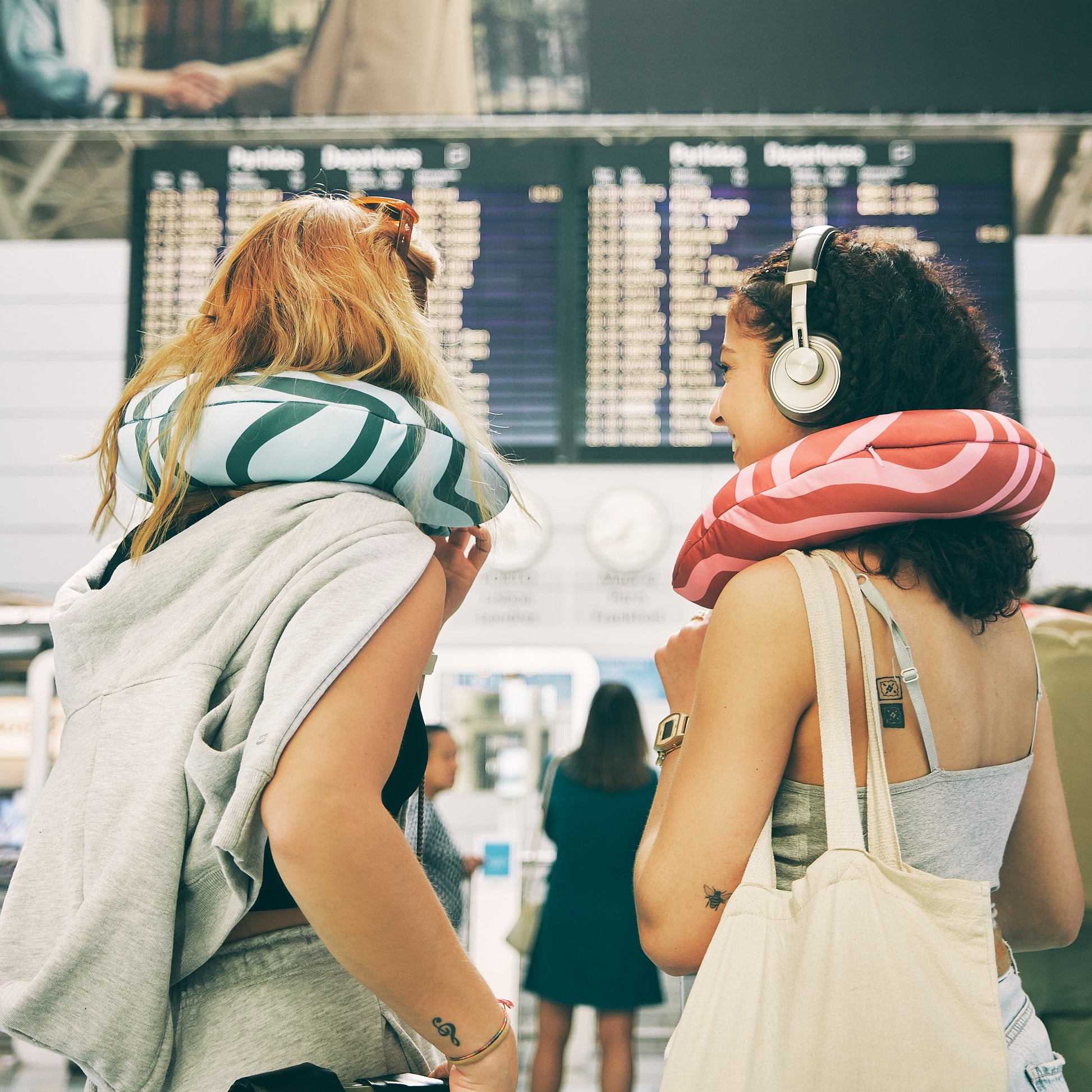 Two travelers wearing blue-green and red-pink neck pillows while waiting at the airport with luggage.