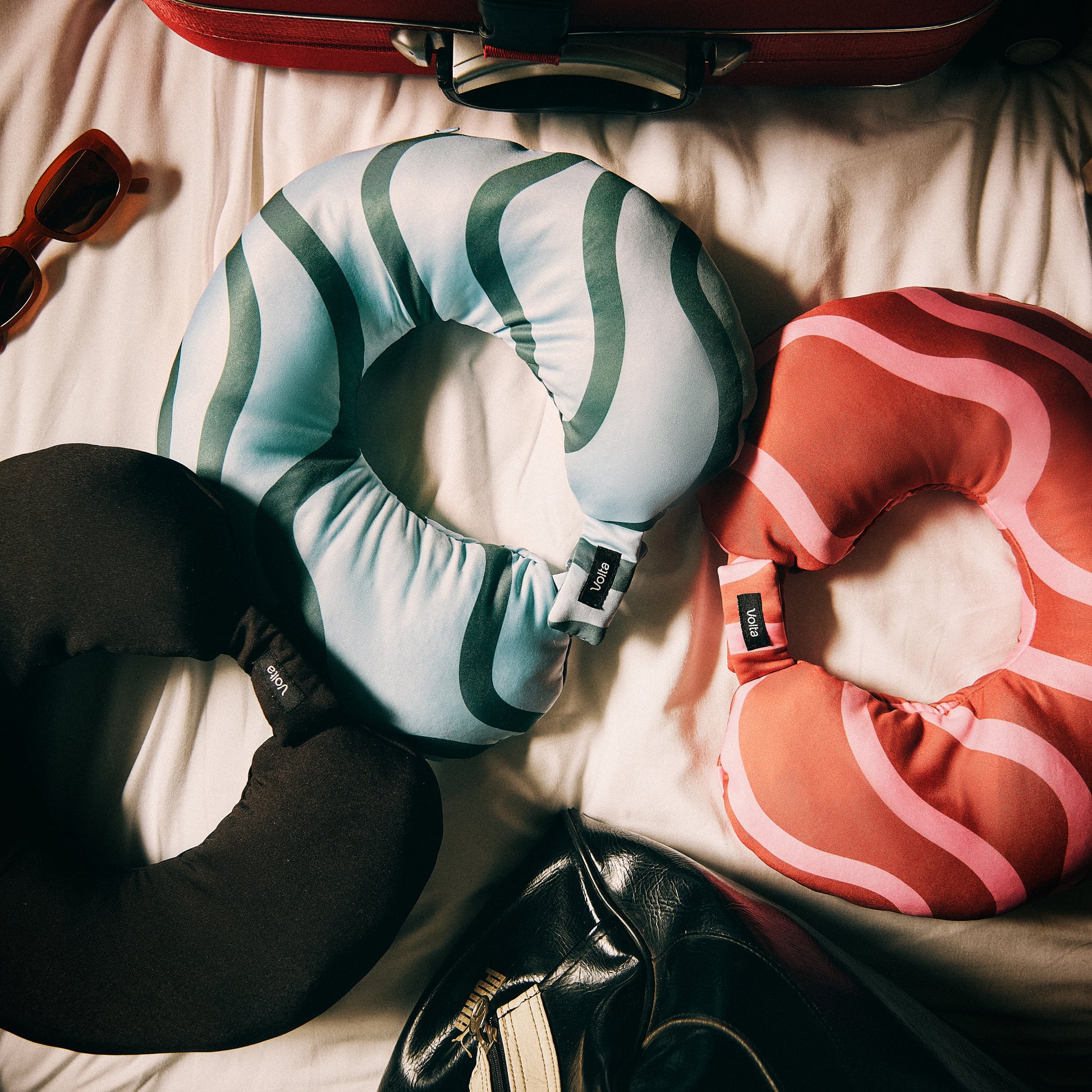 Three travel neck pillows in black, blue-green, and red-pink designs displayed on a bed with luggage and accessories.